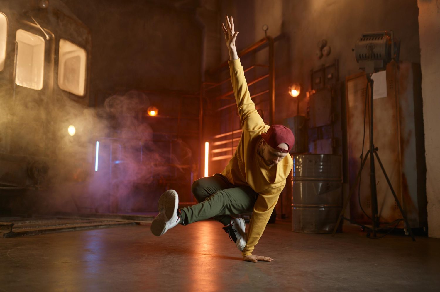 Tancerz uliczny w żółtej bluzie i bordowej czapce wykonuje dynamiczną figurę breakdance opierając się na jednej ręce w industrialnym, zadymionym wnętrzu z kolorowymi światłami w tle.